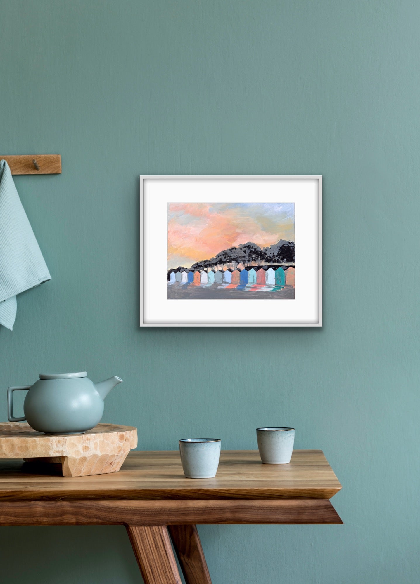 Framed artwork of beach huts on a teal wall above a wooden table with teapots.