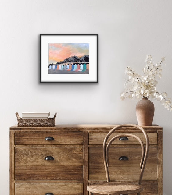 Framed artwork above a wooden dresser with a chair and decorative vase.