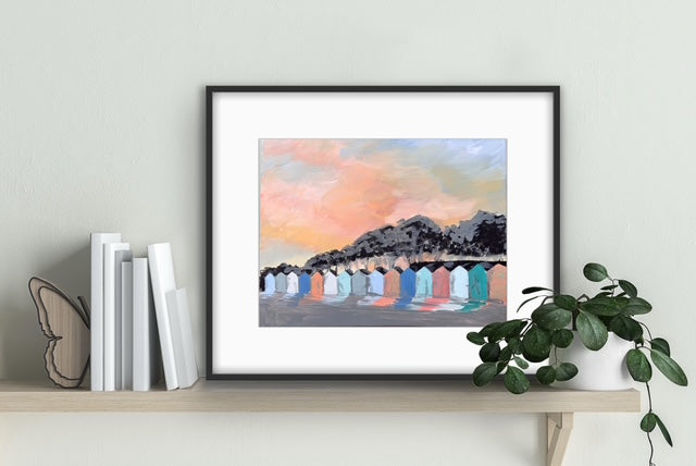 Framed artwork of a mountain landscape with colorful sky on a shelf with books and a plant.